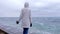 Woman in a white jacket stands on the waterfront on the beach looks at storm waves. Side view.