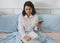 Woman in a white shirt sits on a bed in her bedroom working or studying, talking on the phone and writing