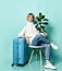Woman in white sweater, jeans, sneakers. Smiling, holding ficus, sitting on chair, leaning on suitcase, posing on blue background
