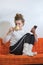 Woman in a white sweater and socks with pompons sits with legs on the sofa and drinks coffee, holding a smartphone