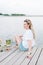 A woman in a white t-shirt sitting in a wooden pier front of water.blondet, smile