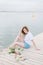 A woman in a white t-shirt sitting in a wooden pier front of water.blondet, smile