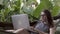 Woman working on laptop while sitting on hammock, exotic background