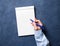 woman writes in notebook on dark blue table, hand in shirt holding a pencil, sketchbook drawing, top view