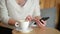 Woman Writting at the Table, Drinking Coffee Or Tea. She Stiring a Cup And Looking At Smartphone. Closeup