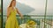 Woman in a yellow dress enjoying a cup of tea, balcony view. Female overlooks a coastal town, reflecting a relaxed