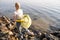 A woman in yellow gloves with a blue garbage bag stands on the rocky shore of the lake. She single-handedly clears the