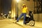 Woman in yellow hat, blouse and blue classic trousers with a black moped - scooter on a night city street