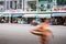 Woman in yellow shirt and red helmet sits on parked motorcycle in front of typical shops and cafes while blurred image of woman