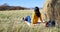 Woman in yellow sweater and rubber boots sitting on plaid next to backpack and thermos with hot tea near haystack in field. she lo