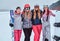 Women friends in sports winter clothes with snowboards and skis standing together in a hug and looking at the camera at