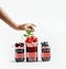 Women hand and jam jars with empty label and various berry fruits at white background. Front view with copy space