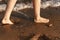 women& x27;s feet washed by a wave walk on a sandy beach.