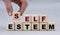 Women`s hand folds wooden cubes with the words SELF ESTEEM on a gray background