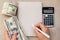 Women`s hand with pen, empty notebook, dollars banknotes and calculator on light wooden background.