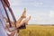 Women`s legs stick out of the car window, against the background of a wheat field.