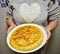 women showing on a white plate a spanish omelet cooked with eggs and potatoes. The woman wears a shirt with a big white heart