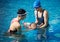 Women in swimming cap and goggles, standing in shallow pool leaning over little child during teaching swimming on back