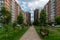 Women walk along a tiled sidewalk past brick multi-storey apartment buildings in a neighborhood on a cloudy summer day