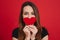 Wonderful happy woman 20s with long brown hair holding paper heart in valentine day at her mouth, isolated over red background