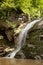 Wonderful summer waterfall in mountain with lush green forest, layered rocks overgrown moss and bright sunbeams on mountain river.