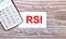 On a wooden background, a white calculator, white paper clips and a white card with the text RSI Relative Strength Index. View