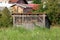 Wooden backyard shed with partially open part filled with hay bales surrounded with high uncut grass and houses in background