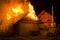 Wooden barn burning at night. High orange fire flames, dense smoke from under tiled roof on dark sky, trees silhouettes and reside