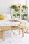 A wooden bedside table with a book and a cup in front of a bed in a natural bedroom interior with many green plants in pots. Real