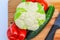 On a wooden Board, fresh vegetables: cauliflower, one red bell pepper, two red tomatoes and one green cucumber. Two sharp ceramic