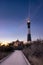 Wooden boardwalk leading to a lighthouse emitting rays of light during blue hour.