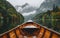 A wooden boat cuts through glassy waters on a misty morning, with lush forested mountains rising dramatically in the