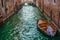 wooden boat floating on the water of a venetian channel between high buildings and a bridge at the end of the channel with some