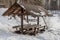 A wooden brown pavilion with table and benches with white snow on the roof in winter in pines and birches forest with a pond