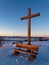 Wooden cross and bench on hill in snow-covered countryside by sunset light