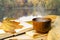 Wooden cup with hot drink, yellow knitted scarf, open book with yellow leaf on background of autumn park with pond.