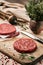 On a wooden cutting board on kraft paper there are raw beef burgers for burgers, spices, rosemary, a pepper mill, a