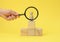 Wooden figurine of a man on a pedestal and a hand with a magnifying glass on a yellow background. The concept of recruiting and