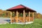 Wooden gazebo structure with new roof and gutter mounted on concrete foundation next to grass and gravel parking with dense trees