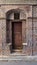 Wooden grunge decorated arched gate with inner small door on wall with black and red bricks