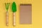 Wooden handled garden hand trowel and hand fork and empty noteboook on yellow background