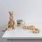 Wooden hare on hinges sits on the edge of  table. Yellow cup with coffee inscription and wooden beads on a white chest of