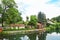 A wooden house between big old trees at the canal embankment, blue pines and a canoe near the wooden fence, a countryside landscap