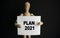 The wooden man holds a white sign with the text PLAN 2021 in his hands. The content of the lettering has implications for business