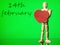 Wooden mannequin with red heart on green color background and 14th February blurred sing in the background, Saint Valentine theme