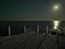 A wooden pier extending into the sea against a dark sky with the moon and a moon path