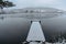 Wooden pier on lake covered with fresh snow.Winter pond with small jetty on misty morning.Foggy cloudy landscape reflected in