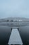 Wooden pier on lake covered with fresh snow.Winter pond with small jetty on misty morning.Foggy cloudy landscape reflected in