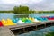 A wooden pier near lakeshore with the row of colored plastic catamarans with lake water and forest on the background