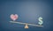 A wooden seesaw on blue chalkboard background with a drawn USD sign overweighing a heart sign